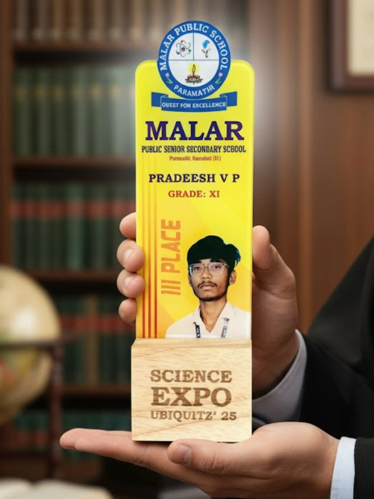 Person holding a trophy with 'Malar Public School' and 'Science Expo Ubiquitz '25' text