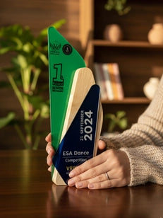 Person holding a 2024 ESA Dance Competition trophy on a wooden table with plants and books in the background.