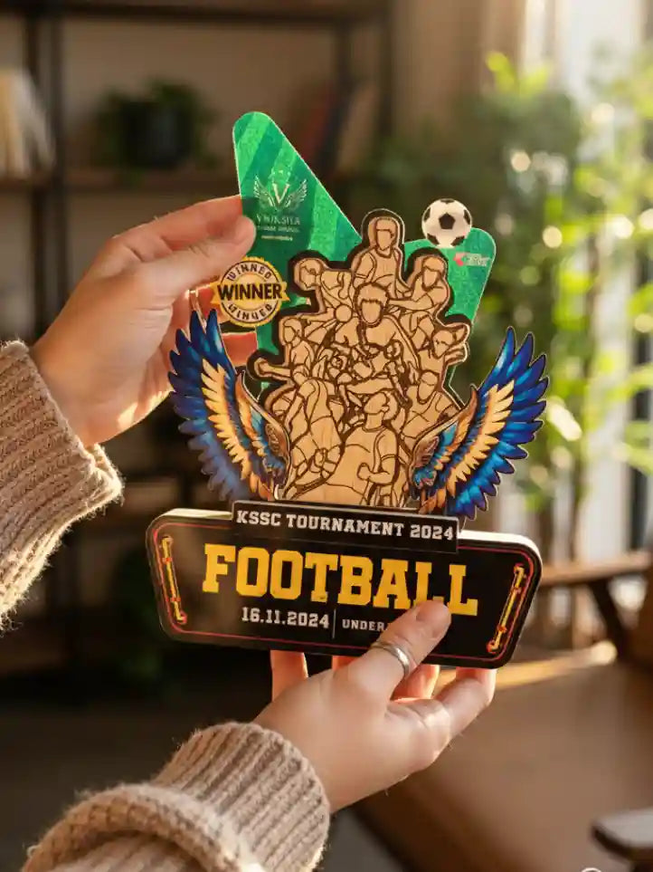 Hand holding a wooden trophy with 'KSSC Tournament 2024 Football' design, blurred indoor background