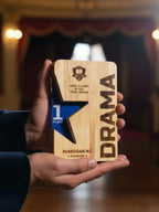 Wooden award with star design and 'Drama' text held in a hand against a blurred theater background