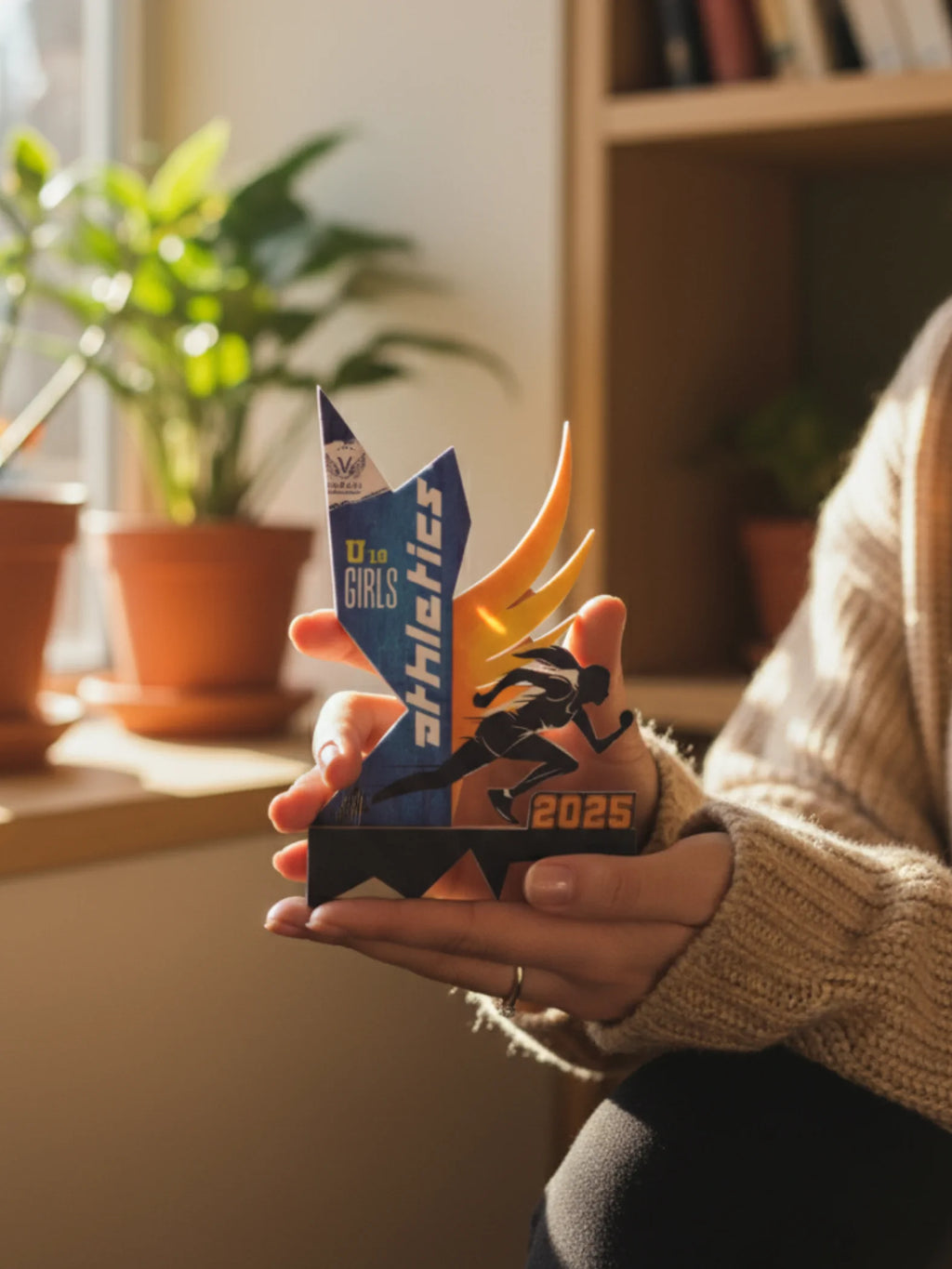 Person holding a colorful trophy with 'Girls Athletics' and '2025' text in a home setting.
