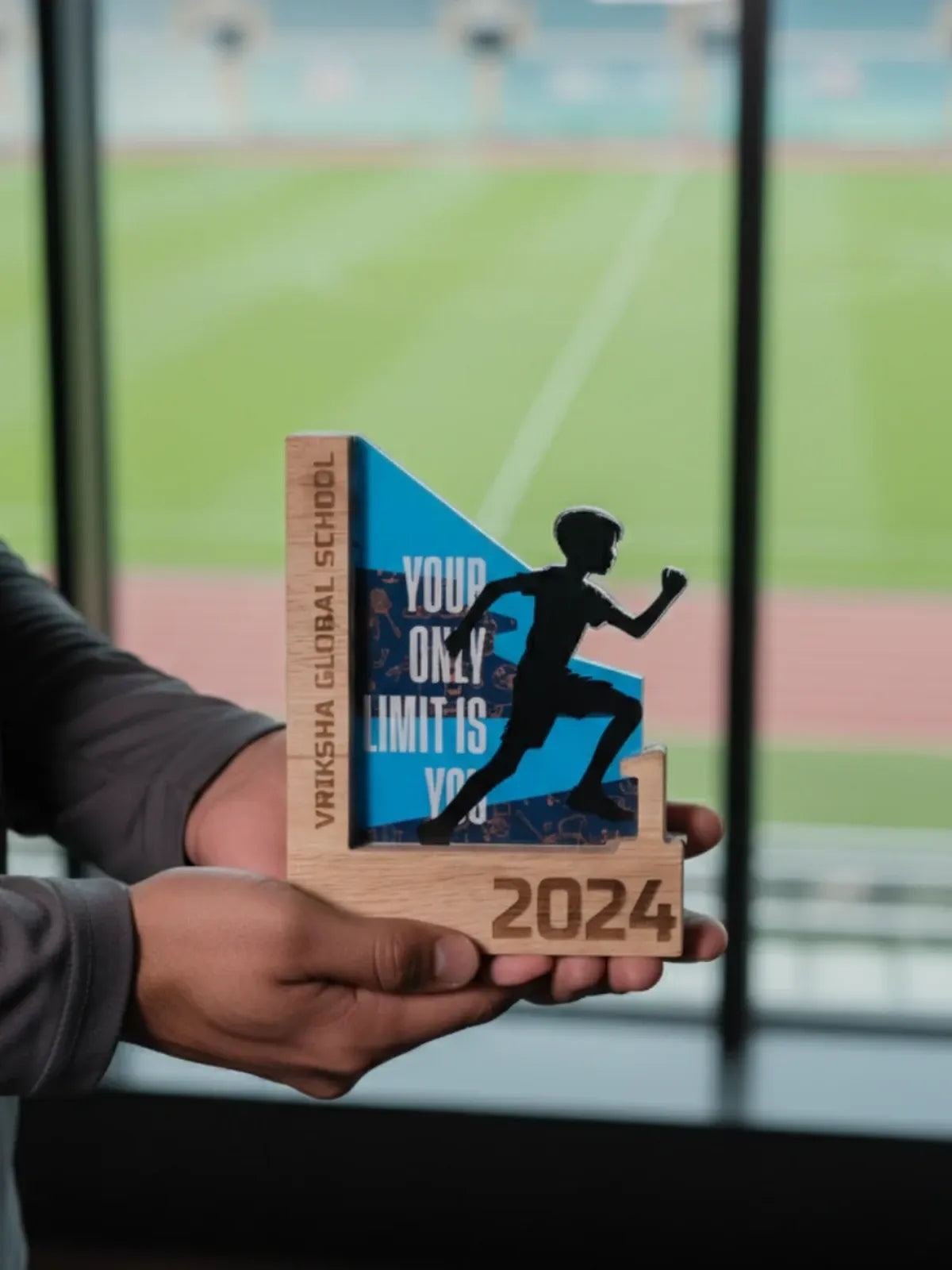 Wooden trophy with a runner silhouette and 'Vrishna Global School' text, held by a person in front of a sports field.