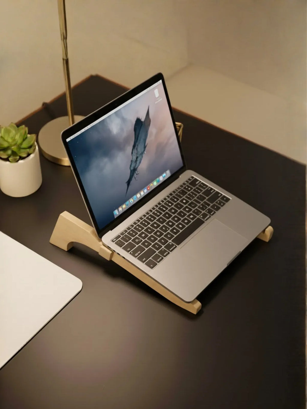 Laptop on a wooden stand on a desk with a plant and lamp in the background