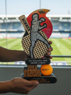 Table tennis trophy with a paddle design held by a person, set against a sports venue background.