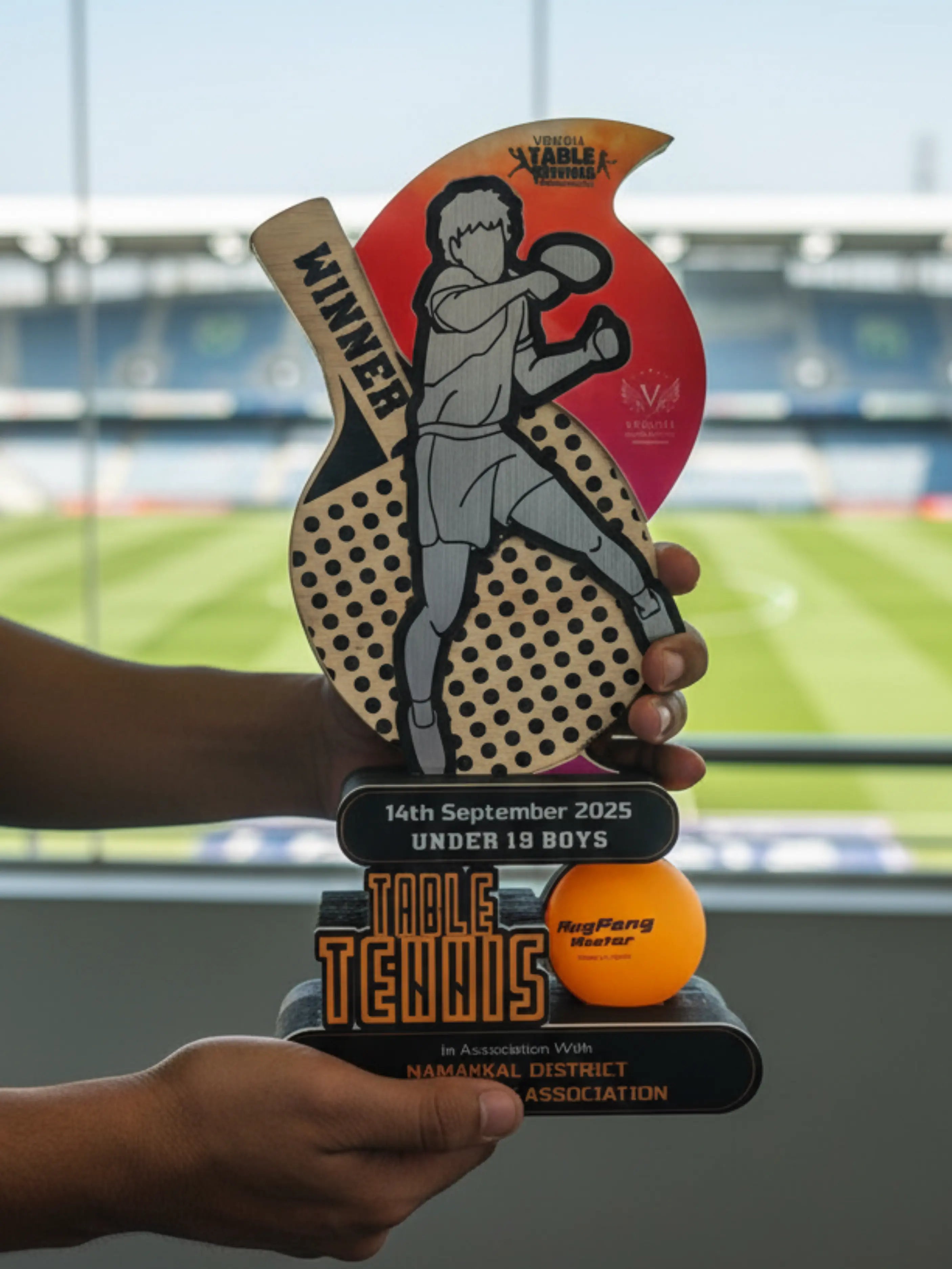 Table tennis trophy with a paddle design held by a person, set against a sports venue background.