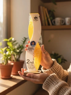 Person holding a bottle of 2nd Edition juice in a home setting with plants and books.