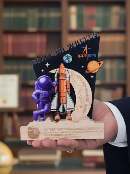 A person holding a space-themed trophy with an astronaut figure, rocket, and celestial design in front of a bookshelf.