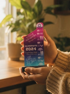 Hand holding a colorful Olympic 2024 badge with plants and a window in the background