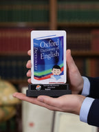 Hand holding a miniature Oxford Dictionary of English against a blurred bookshelf background