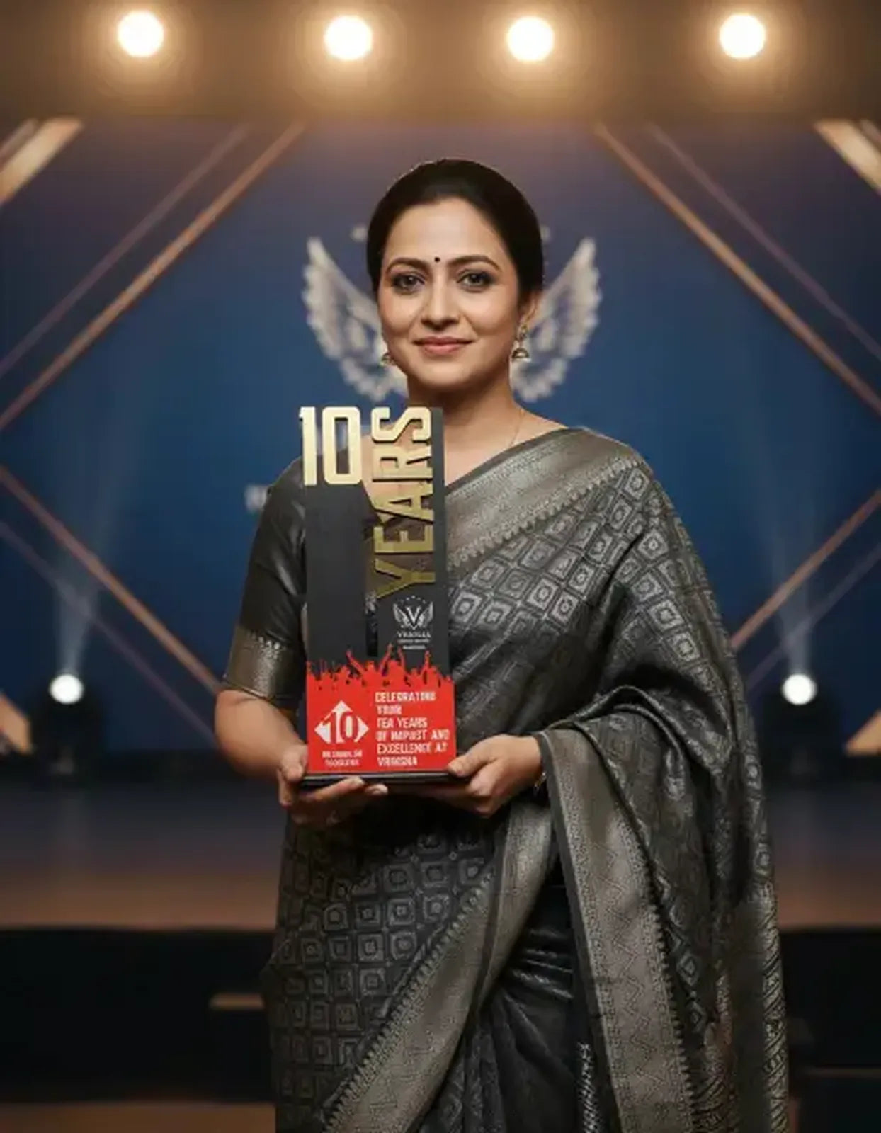 Woman in a saree holding an award with '10 Years' on it, against a stage background.