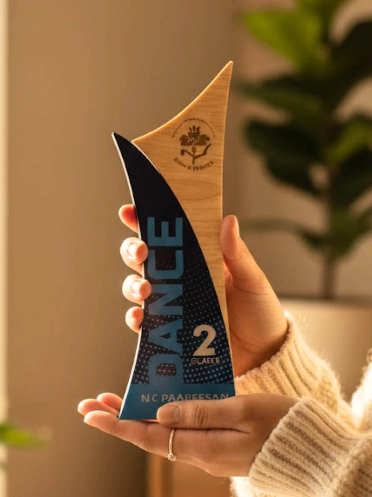 Person holding a decorative award with 'DANCE' and 'N.C. PAARESS' text, blurred indoor background